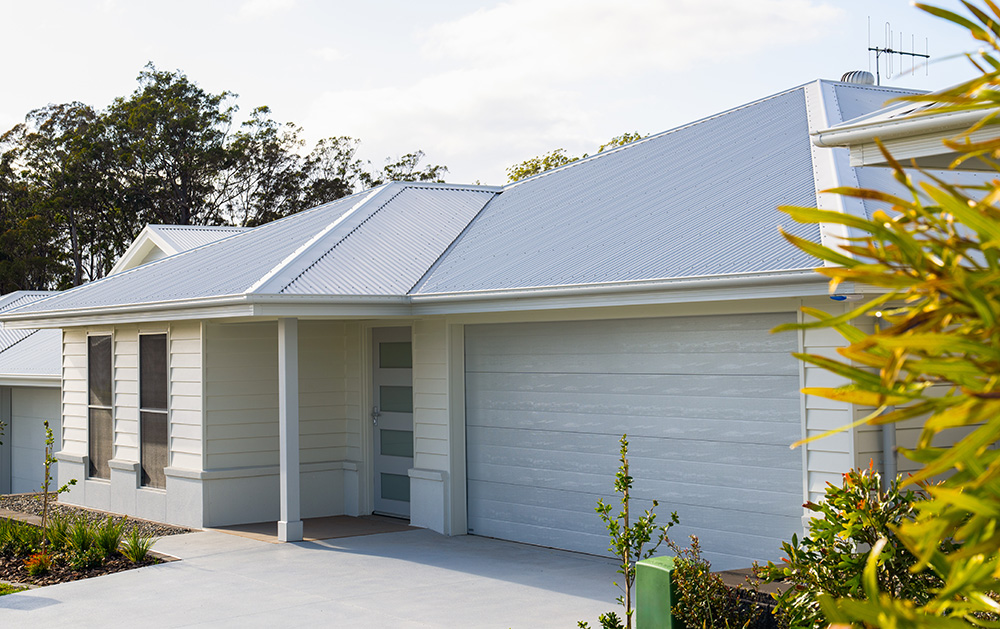 COLORBOND steel cool roofing.Single-story house with light grey cladding and a steel roof, featuring a garage and small porch. Surrounded by lush greenery under a partly cloudy sky.