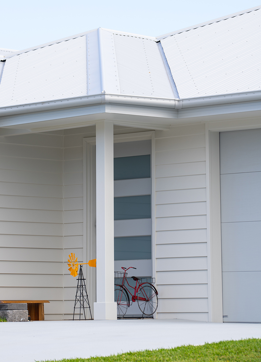 COLORBOND steel cool roofing. Modern house front porch with white cladding and a white steel roof. A red bicycle with baskets leans against a wall, next to a small decorative windmill.